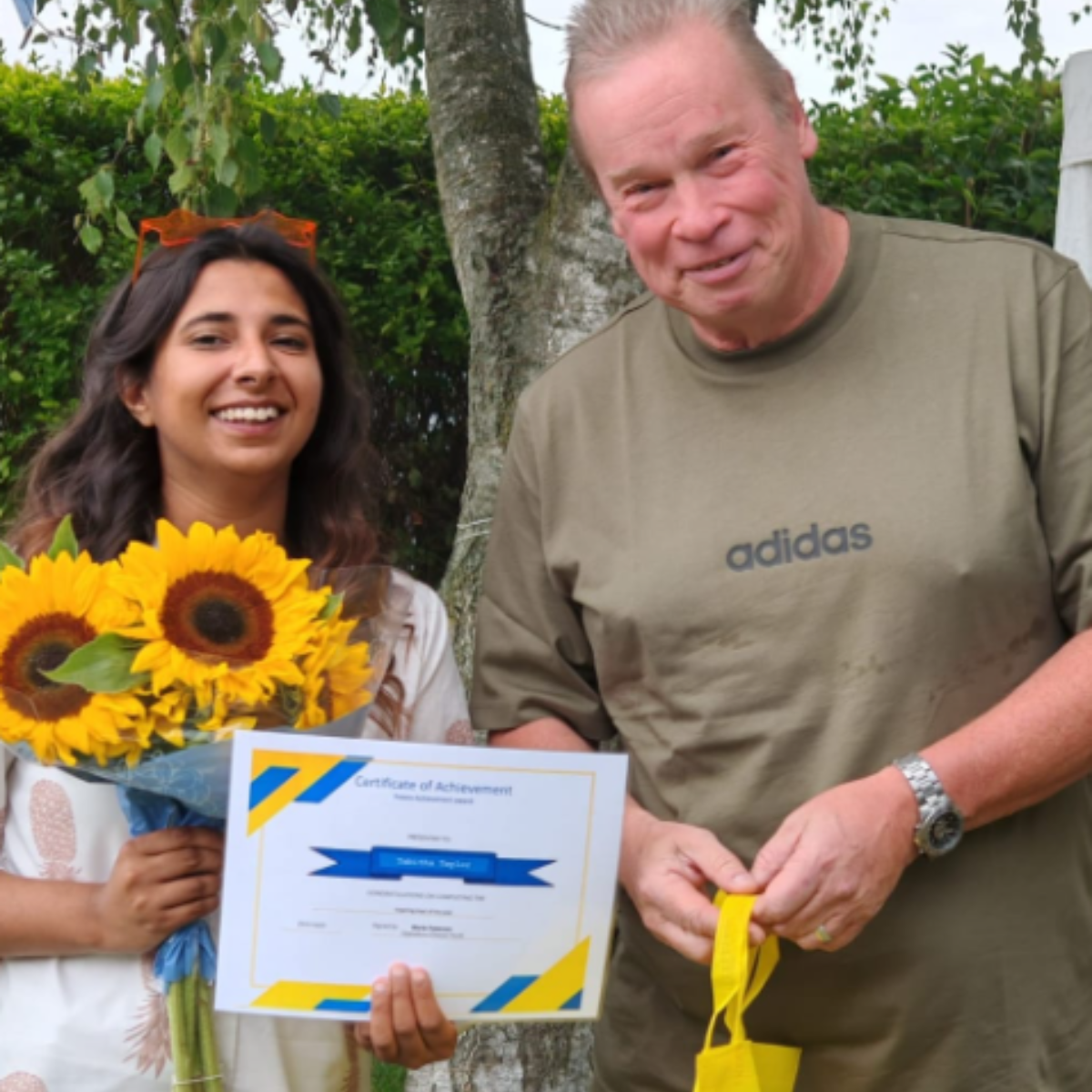 A woman with sunflowers and a certificate of achievement smiles next to a man wearing an Adidas shirt.