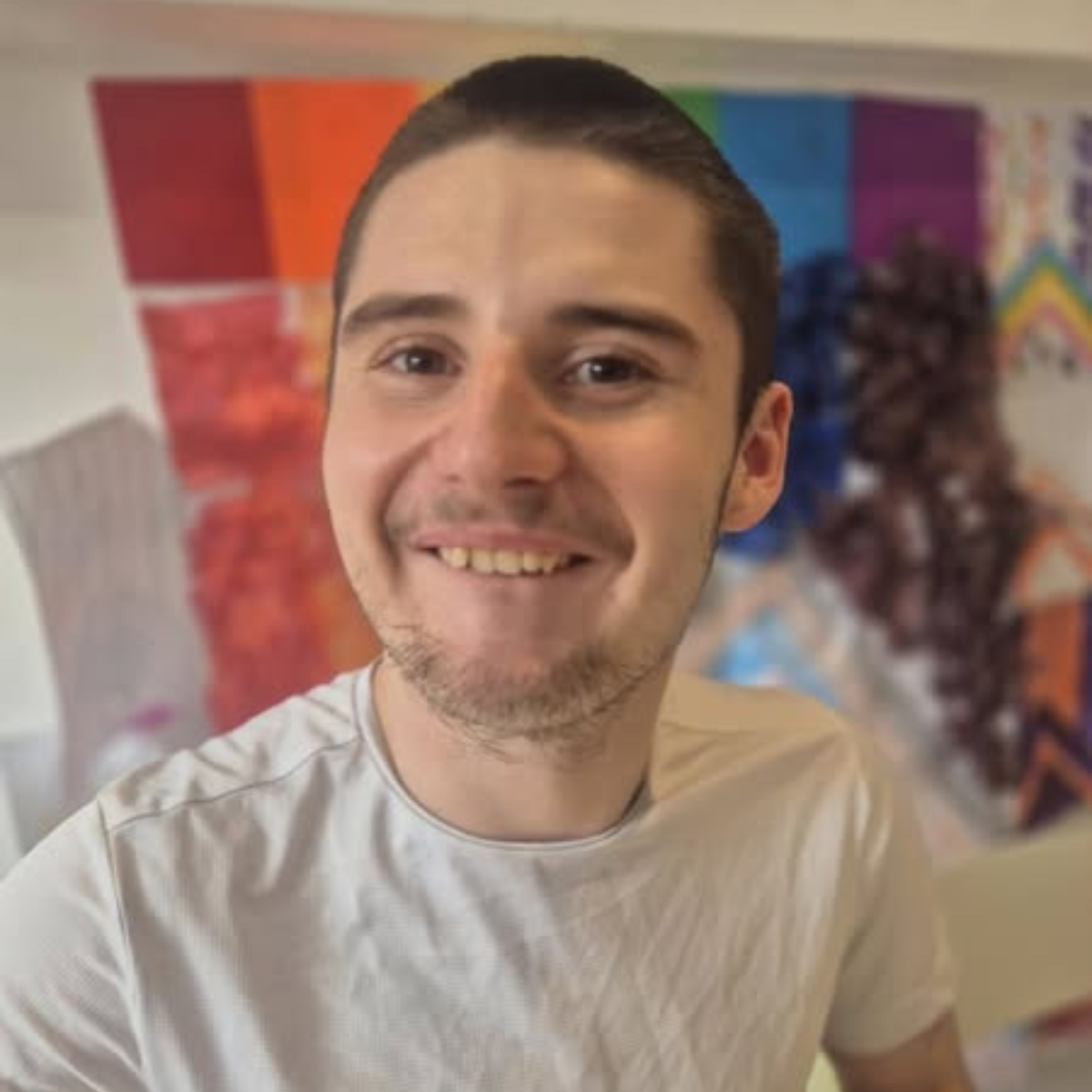 A young man with short brown hair and stubble smiles at the camera. He wears a light gray t-shirt. Behind him, a wall features colorful, abstract art and a rainbow stripe.