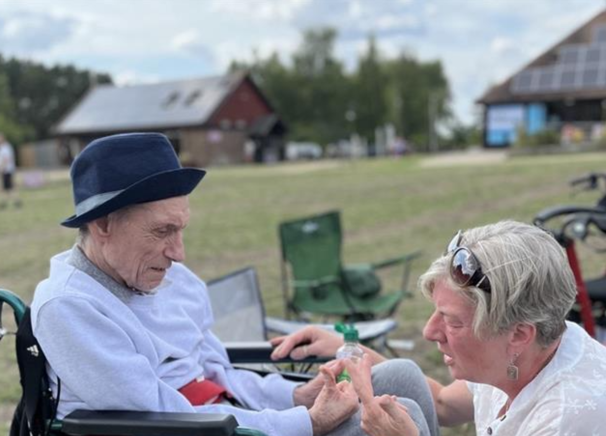 A woman in a white shirt and sunglasses helps an elderly man in a wheelchair, possibly with medication or a small item, outdoors with buildings in the background.