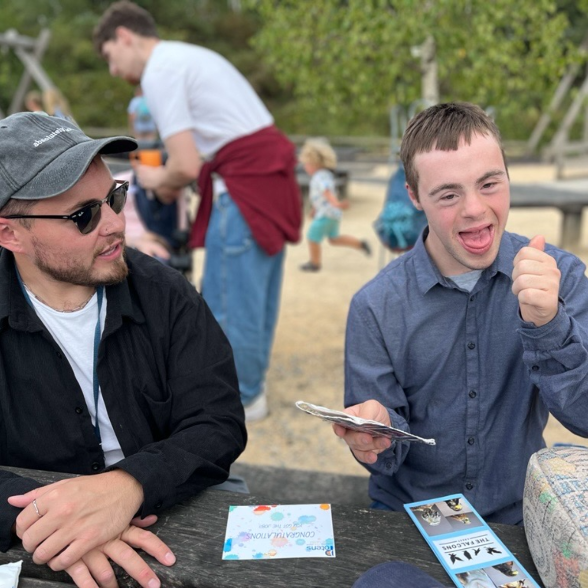 A young man with Down syndrome gives a thumbs up while holding papers. Another young man sits nearby.