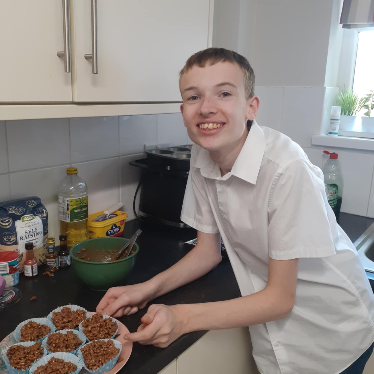 A young person proudly displays homemade chocolate Rice Krispie treats in a kitchen setting, with ingredients and a mixing bowl nearby.