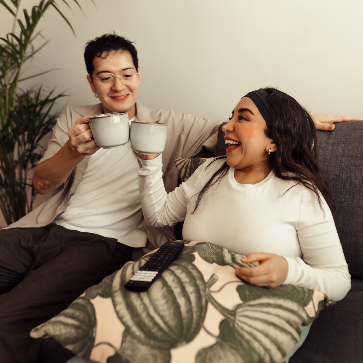 A smiling couple raises coffee mugs while sitting on a sofa.