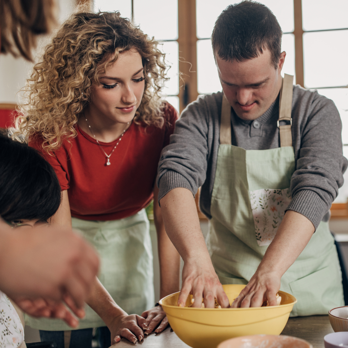 A man with Down syndrome and a woman with curly hair, both wearing aprons, knead dough in a yellow bowl.