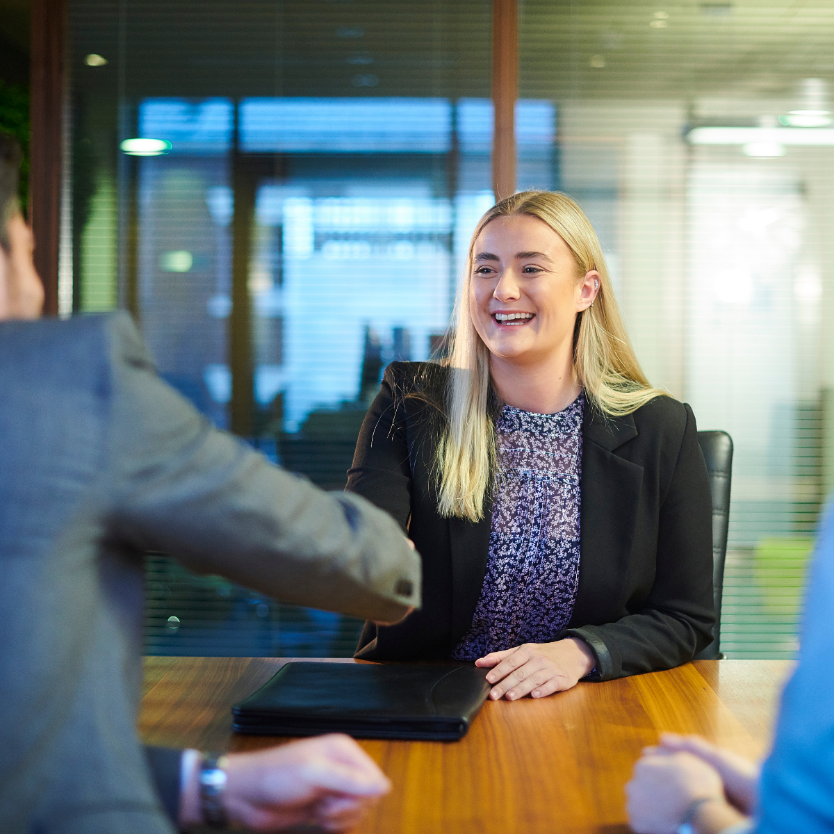 A smiling woman shakes hands with an interviewer across a desk.