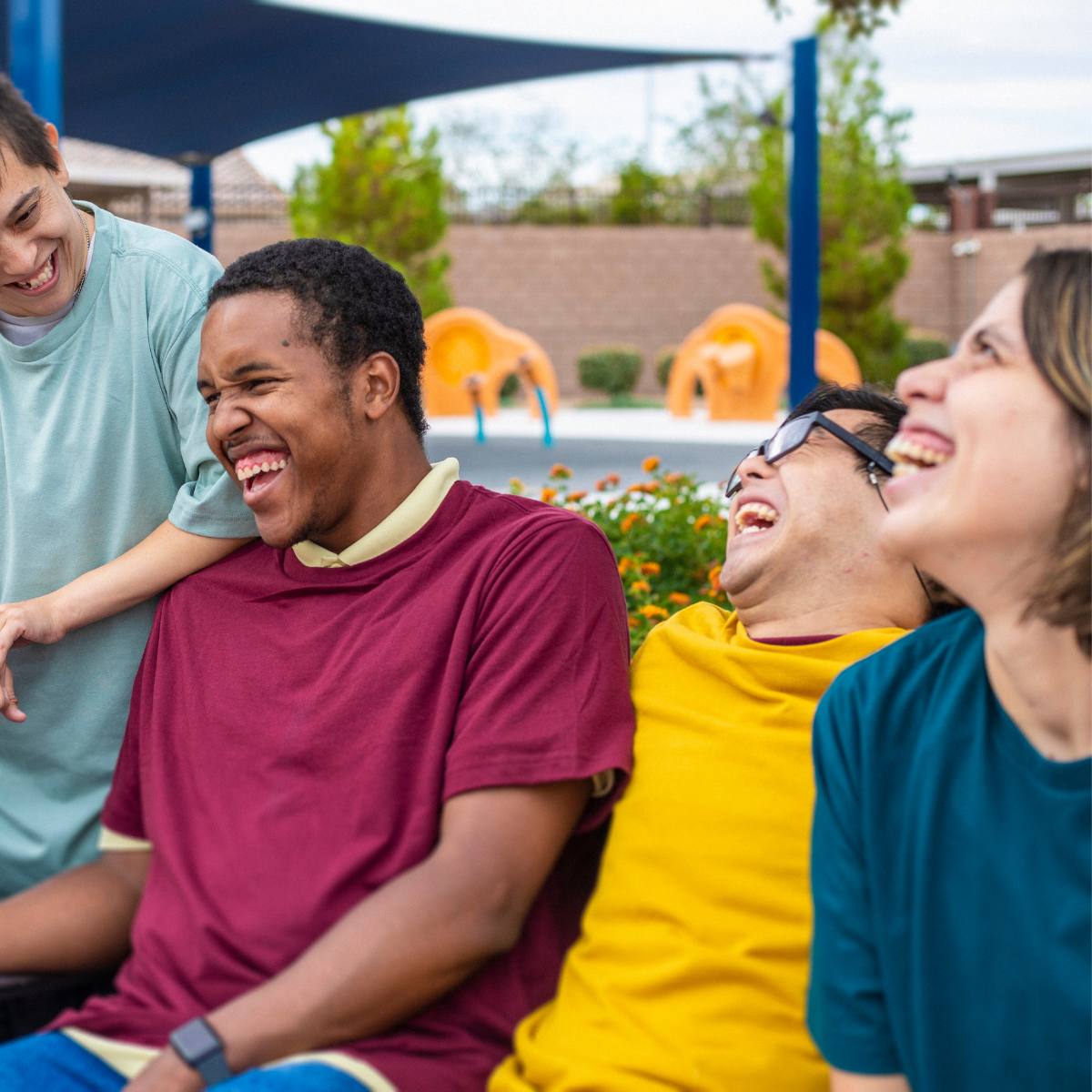 Four friends with diverse abilities laugh together outdoors at a park, enjoying a sunny day.