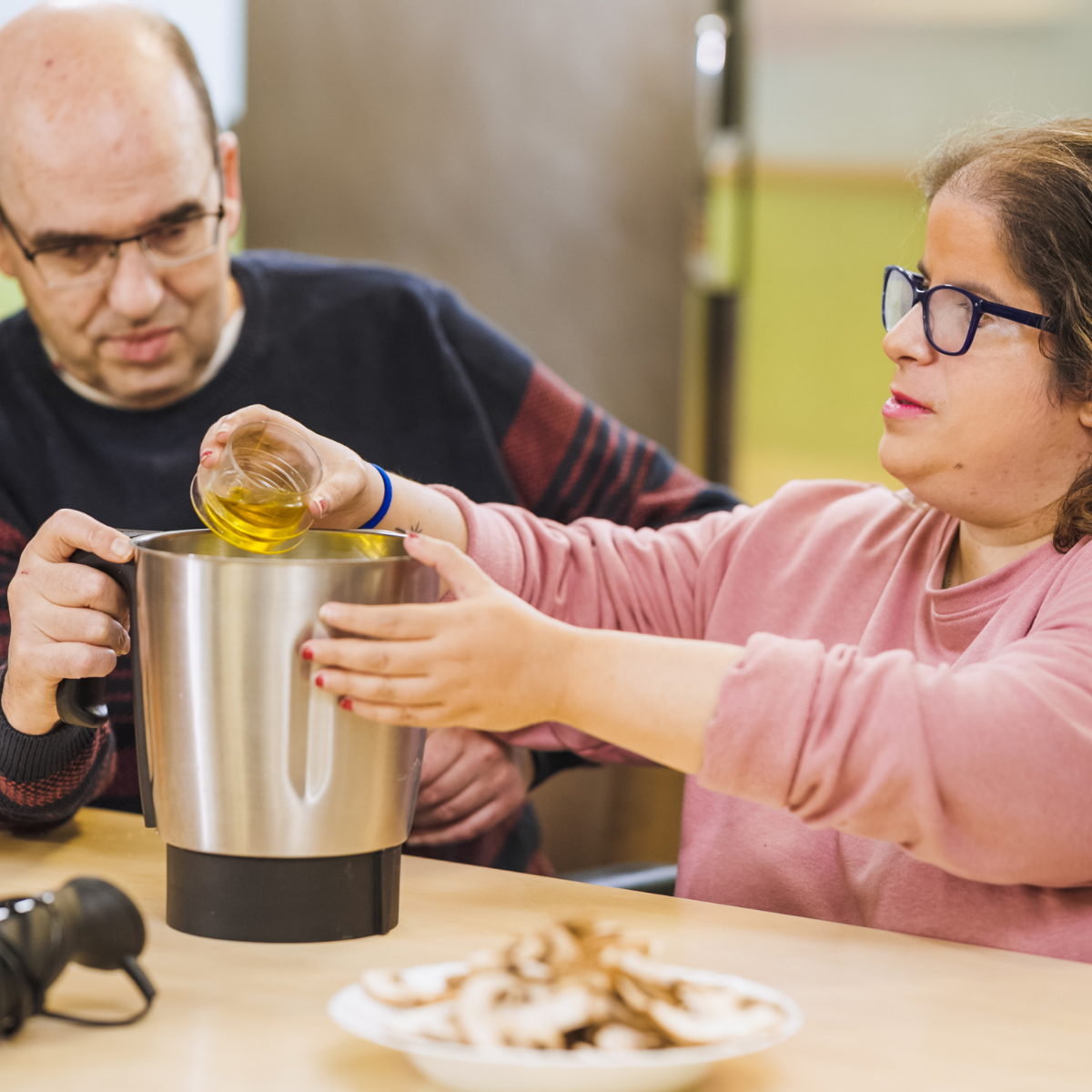 A woman pours oil into a food processor while a man watches. Sliced mushrooms are on a plate in the foreground.