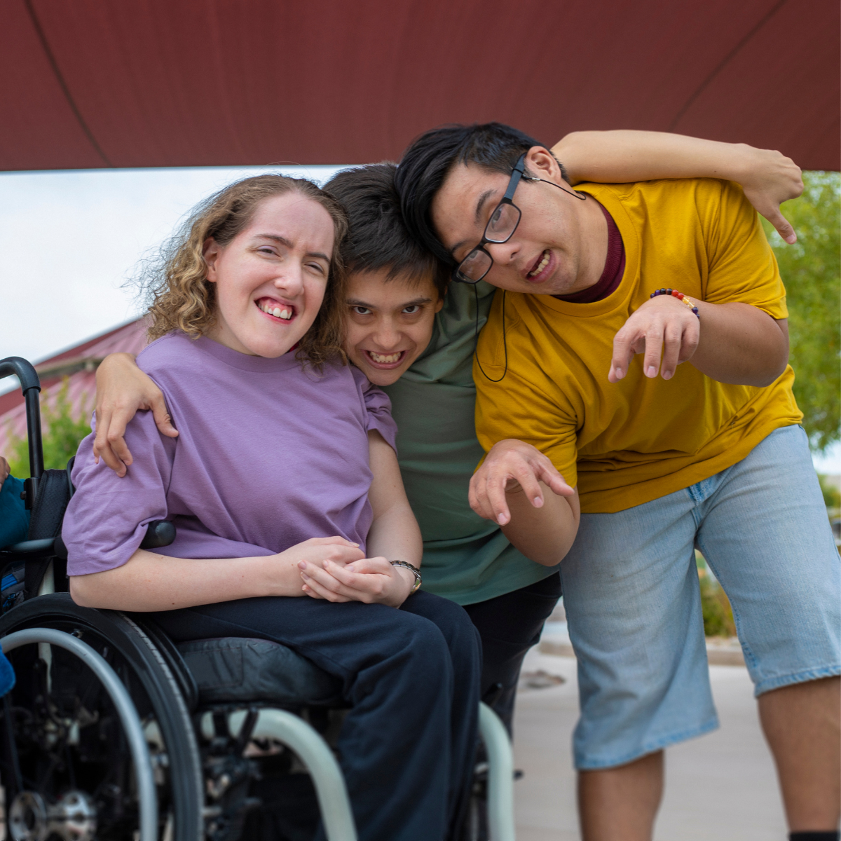 Three young friends, one in a wheelchair, are hugging and smiling at the camera outdoors.