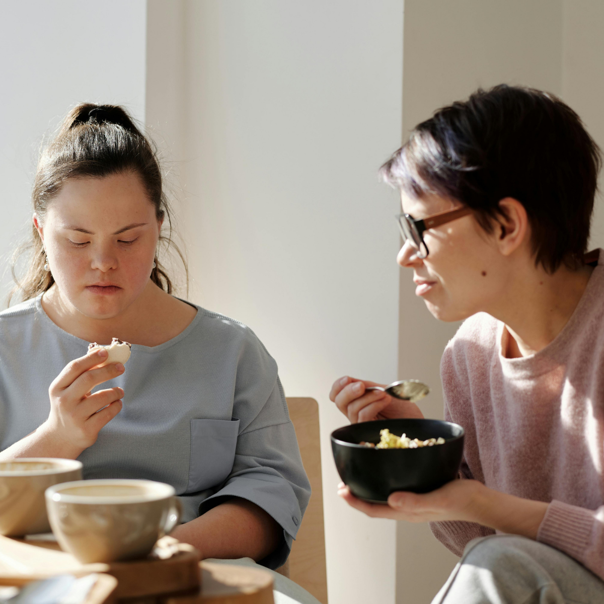 A woman with Down syndrome eats a pastry while another woman eats from a bowl.