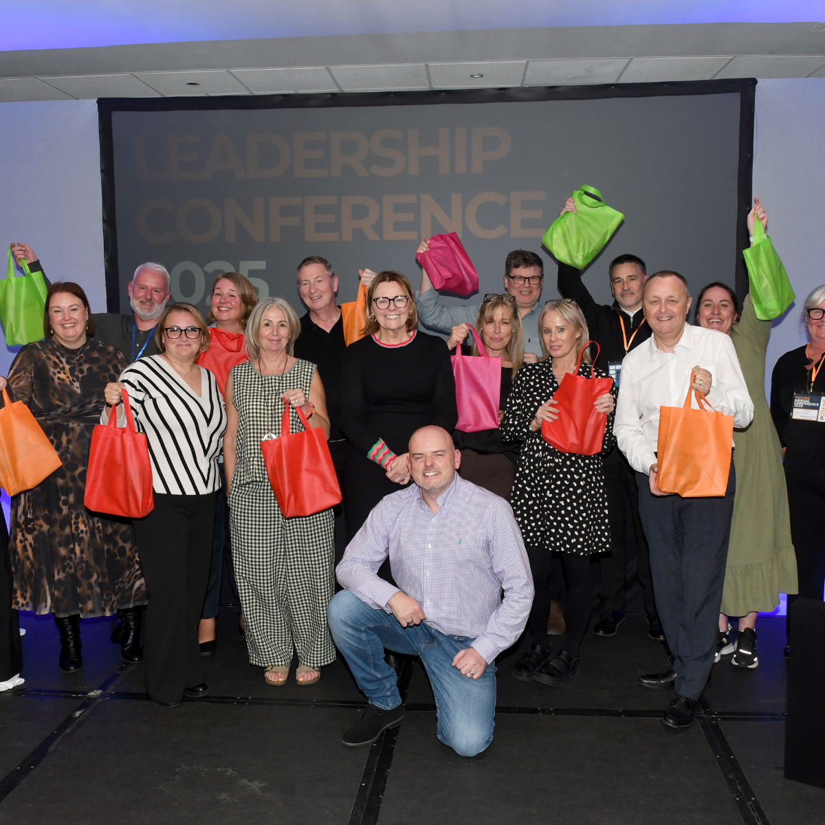 Attendees at a conference pose for a group photo, holding gift bags of various colors. The screen behind them displays "LEADERSHIP CONFERENCE 2023."
